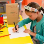 A child sits at a bright yellow craft table, drawing on a sheet of white paper with a black marker. The table is scattered with colourful craft supplies, including felt‑tip pens, scissors, paper shapes, and a cardboard model decorated with crayon drawings. The child wears a patterned headband and a blue cardigan, with braided hair hanging over one shoulder. The room has orange walls and large windows in the background.