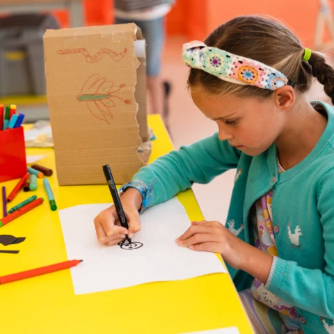 A child sits at a bright yellow craft table, drawing on a sheet of white paper with a black marker. The table is scattered with colourful craft supplies, including felt‑tip pens, scissors, paper shapes, and a cardboard model decorated with crayon drawings. The child wears a patterned headband and a blue cardigan, with braided hair hanging over one shoulder. The room has orange walls and large windows in the background.