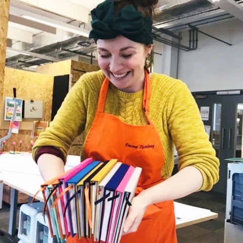 A person wearing an orange apron over a yellow knitted sweater stands in a workshop space, holding a bundle of brightly coloured handmade notebooks. The notebooks are stacked together with their covers facing outward and ribbon-like ties hanging from the spines. The workshop environment includes tables, tools, and storage units in the background.