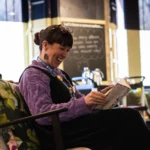A person sits in a cushioned armchair inside a café or lounge setting, holding and reading an open book. The chair has patterned cushions, and a small table is nearby. The background features a counter area with metal taps, a chalkboard-style menu board with handwritten text, and tall windows with light filtering through. The space has warm indoor lighting and a relaxed seating arrangement.