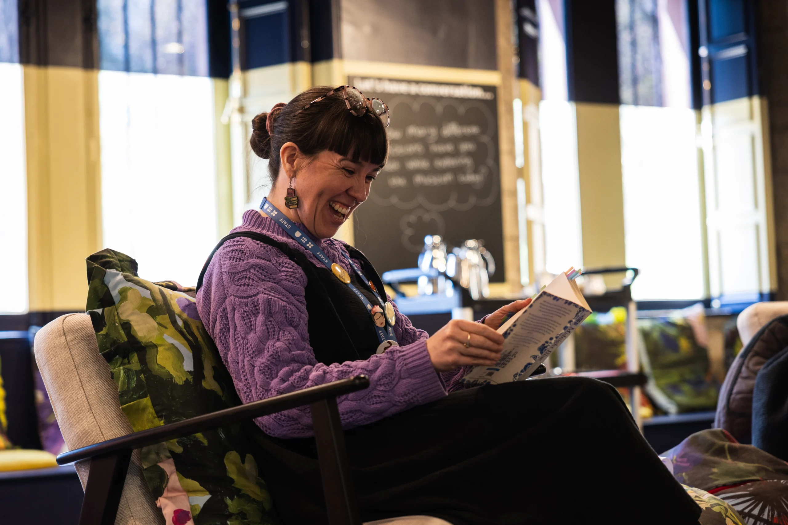 A person sits in a cushioned armchair inside a café or lounge setting, holding and reading an open book. The chair has patterned cushions, and a small table is nearby. The background features a counter area with metal taps, a chalkboard-style menu board with handwritten text, and tall windows with light filtering through. The space has warm indoor lighting and a relaxed seating arrangement.
