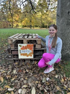 A child crouches beside a small outdoor “bug hotel” made from stacked wooden pallets filled with twigs, leaves, and natural materials. A hand‑painted sign on the front reads “BUG HOTEL” and is decorated with autumn leaves and a small illustrated insect. The child wears a long‑sleeved grey top, bright pink trousers, and trainers, and rests one hand on the structure. The setting is a park or garden with grass, fallen leaves, trees with autumn foliage, and a large tree trunk behind the bug hotel.