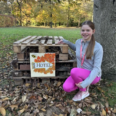 A child crouches beside a small outdoor “bug hotel” made from stacked wooden pallets filled with twigs, leaves, and natural materials. A hand‑painted sign on the front reads “BUG HOTEL” and is decorated with autumn leaves and a small illustrated insect. The child wears a long‑sleeved grey top, bright pink trousers, and trainers, and rests one hand on the structure. The setting is a park or garden with grass, fallen leaves, trees with autumn foliage, and a large tree trunk behind the bug hotel.