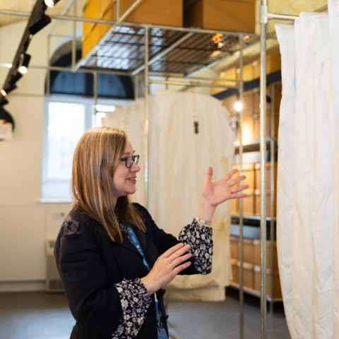 A person stands inside a storage or project space with metal shelving and suspended fabric panels. The person gestures with one hand toward a tall white fabric surface hanging from a metal structure. The surrounding area includes stacked boxes, shelving units, and overhead track lighting, with daylight visible through a window in the background.