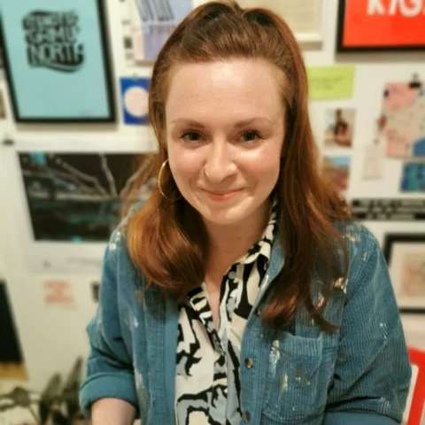 A person stands indoors in front of a wall covered with framed artwork and prints. They are wearing a patterned shirt under a blue, paint‑spattered overshirt. Their hair is shoulder‑length and auburn. A plant is partially visible in the lower corner of the scene.