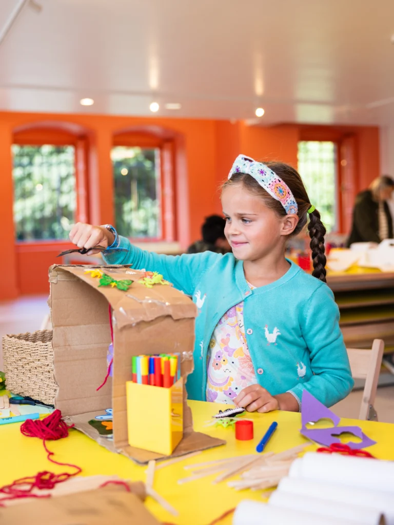 Photograph of a child engaged in a craft activity at a brightly lit classroom with orange walls and large windows. The child is decorating a cardboard structure with various craft supplies, including markers, yarn, and paper shapes, highlighting a creative and educational environment.