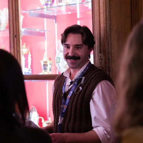 A person wearing a textured brown knitted vest over a white shirt stands inside a dimly lit gallery space. Glass display cabinets behind them contain decorative objects, including ornate ceramics and figurines, illuminated against a vibrant red backdrop. Two other visitors are visible in the foreground, facing the person who appears to be speaking or guiding a discussion.