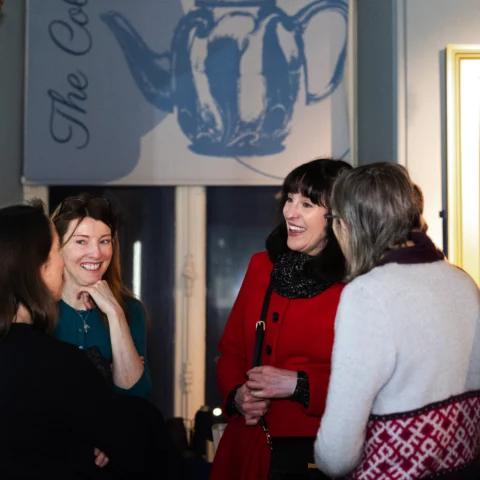 A small group of people stand together in a softly lit exhibition space, engaged in conversation. They are surrounded by display cases and framed artwork, including a large blue illustration of a teapot hanging behind them. One individual wears a bright red coat, while another has a patterned knit garment. The setting appears to be a museum or gallery during an event or gathering.