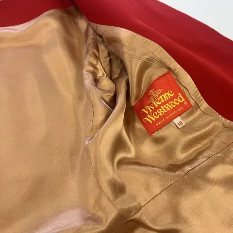 The interior of a red garment is shown with the lining pulled open to reveal a gold satin interior. A bright red fabric label stitched into the lining reads “Vivienne Westwood” in yellow lettering, along with a small size tag marked “10.” The outer red fabric of the garment is visible at the edge of the image.