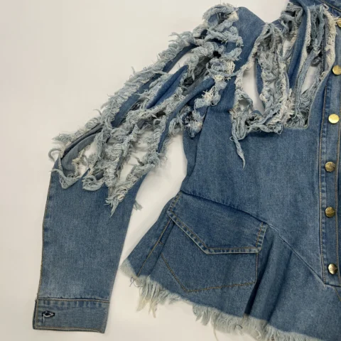 A denim jacket is shown laid flat on a light background. The jacket features heavily distressed, shredded detailing along the upper sleeve and shoulder area, creating long frayed strips of denim. The rest of the sleeve is intact, while the lower hem of the jacket has a frayed, unfinished edge. Brass‑coloured buttons run down the front, and a large, angled pocket is visible near the waist.