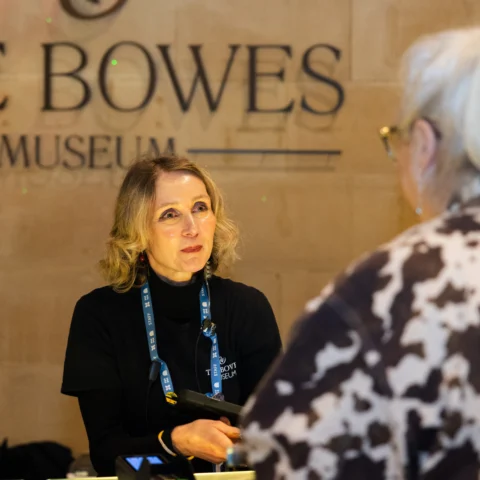 A staff member sits behind an information or reception desk inside The Bowes Museum, identifiable by the large logo on the wall behind them. Another visitor stands on the opposite side of the desk, and the staff member appears to be assisting them. Various lanyards and equipment are visible on the desk.
