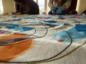 A large piece of fabric lies stretched across a table, covered in bold abstract artwork featuring sweeping black lines and areas of bright colour including orange, blue, purple, and pink. Two people stand at the far side of the table working on the fabric—one wearing gloves and applying paint, and the other leaning in while holding a tool or brush. The scene is set in a well‑lit room with tall windows in the background.