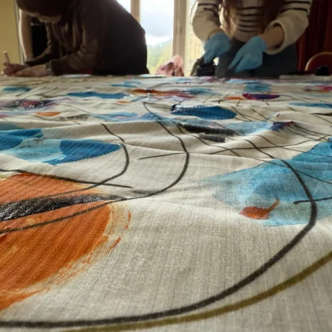 A large piece of fabric lies stretched across a table, covered in bold abstract artwork featuring sweeping black lines and areas of bright colour including orange, blue, purple, and pink. Two people stand at the far side of the table working on the fabric—one wearing gloves and applying paint, and the other leaning in while holding a tool or brush. The scene is set in a well‑lit room with tall windows in the background.