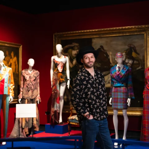 A person stands in the foreground of a fashion exhibition gallery, surrounded by mannequins dressed in vibrant and patterned garments. The display includes tailored suits, dresses, and coordinated outfits in colours such as red, blue, tartan, and multicoloured patchwork. The walls of the gallery are deep red, and a large framed painting hangs behind the mannequins. Exhibition lighting highlights the clothing while the room remains dim overall.