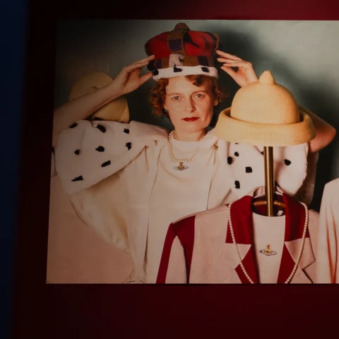 A photograph shows a person wearing a crown and cape posing among hats displayed on stands, printed and mounted on a red gallery wall.