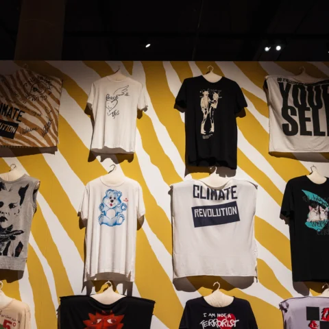A wall display of graphic T‑shirts mounted against a bold yellow‑and‑white striped background. The shirts are arranged in a grid across several rows and feature printed slogans, illustrations, and photographic images in black, white, and muted colours. Visible text on some shirts includes phrases such as “CLIMATE REVOLUTION” and large block lettering reading “YOUR SELF.” The display is lit by gallery spotlights within a dark exhibition space.
