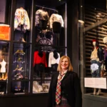 A person stands in the foreground of a fashion exhibition gallery, positioned in front of tall glass display cases. Inside the cases are mannequins dressed in tailored jackets, skirts, and patterned garments, alongside accessories such as shoes and small objects. A bright orange bag with the text “Vivienne Westwood” is visible on an upper shelf to the left. Additional mannequins wearing coordinated outfits stand to the right of the person. The space is dimly lit with display lighting highlighting the clothing and objects against dark walls.