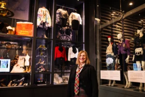 A person stands in the foreground of a fashion exhibition gallery, positioned in front of tall glass display cases. Inside the cases are mannequins dressed in tailored jackets, skirts, and patterned garments, alongside accessories such as shoes and small objects. A bright orange bag with the text “Vivienne Westwood” is visible on an upper shelf to the left. Additional mannequins wearing coordinated outfits stand to the right of the person. The space is dimly lit with display lighting highlighting the clothing and objects against dark walls.