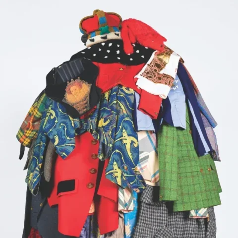 A full‑body figure stands against a plain light background wearing an elaborate sculptural outfit made from many layered garments. The clothing pieces are brightly coloured and vary in patterns and textures, including jackets, shirts, skirts, and fabric panels stacked and draped to form a voluminous, mound‑like silhouette. The figure also wears bright red tights and matching shoes, with only the legs visible beneath the mass of clothing.
