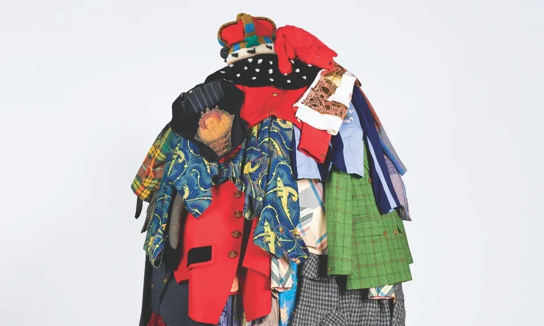 A full‑body figure stands against a plain light background wearing an elaborate sculptural outfit made from many layered garments. The clothing pieces are brightly coloured and vary in patterns and textures, including jackets, shirts, skirts, and fabric panels stacked and draped to form a voluminous, mound‑like silhouette. The figure also wears bright red tights and matching shoes, with only the legs visible beneath the mass of clothing.