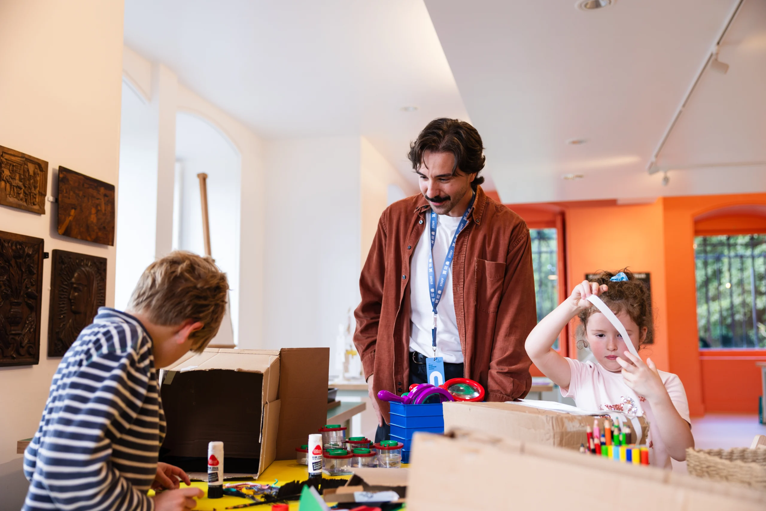 A workshop scene inside a museum or gallery space shows an adult standing behind a table while two children work on craft activities. The table is covered with cardboard boxes, coloured paper, glue sticks, pens, scissors, tape, and small containers of materials. One child bends over the table working inside a cardboard structure, while another holds a strip of white paper above the table. The room has white walls, display panels with carved objects on the left, and bright orange walls and windows in the background, lit by natural and overhead lighting.