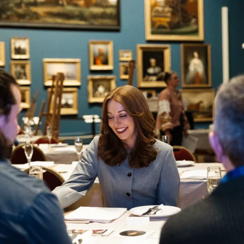 Visitors sit around a table set with plates and drinks inside a museum gallery, with easels and a wall of framed paintings behind them. One person in the centre looks down at materials on the table while others sit nearby in conversation.