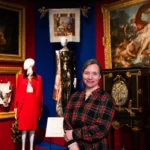 A person stands in the foreground of a fashion exhibition gallery with arms folded. Behind them, mannequins display tailored garments, including a bright red coat and a dark, patterned dress. The gallery walls are painted deep red and blue and feature large framed classical paintings in ornate gilt frames. A richly decorated cabinet with inlaid designs stands to the right, and a small exhibition label is visible near the mannequins. Display lighting highlights the clothing and artworks against the darker surroundings.