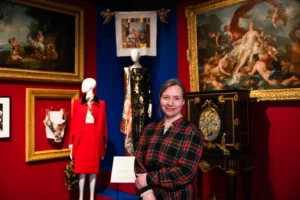 A person stands in the foreground of a fashion exhibition gallery with arms folded. Behind them, mannequins display tailored garments, including a bright red coat and a dark, patterned dress. The gallery walls are painted deep red and blue and feature large framed classical paintings in ornate gilt frames. A richly decorated cabinet with inlaid designs stands to the right, and a small exhibition label is visible near the mannequins. Display lighting highlights the clothing and artworks against the darker surroundings.