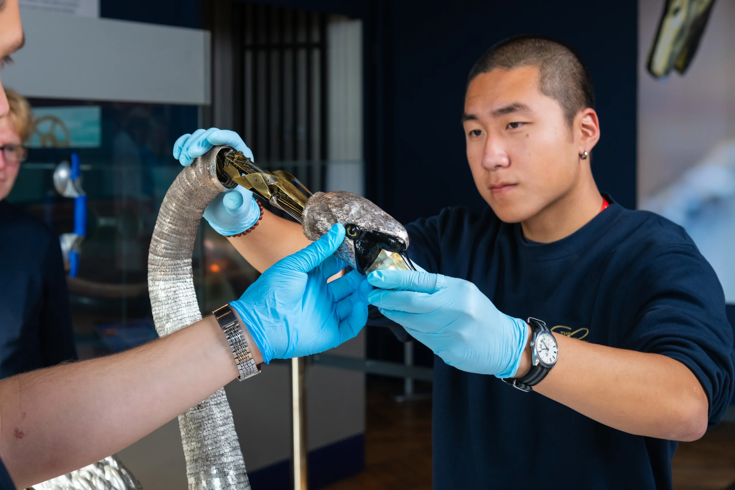 Two people wearing protective gloves carefully handle a mechanical bird object, with its beak and internal mechanism partially exposed, in a museum conservation or workshop setting.