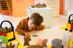 A child sits at a bright yellow table drawing on a large sheet of white paper with a red marker. The child leans forward with one arm resting on the table while drawing a small coloured shape near the centre of the page. Containers filled with coloured pens and pencils sit on the table to both sides. In the foreground are small cardboard cut‑out shapes, and in the background a display plinth holds an object within a well‑lit indoor museum or gallery space.