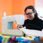 A person sits at a table using a white Singer sewing machine, guiding a piece of white fabric under the needle. The table is covered with colourful fabric pieces in the foreground. The scene is set in a bright indoor workshop space with orange walls and soft natural light coming from windows behind. The sewing machine, fabric, and tools are the main focus of the image.