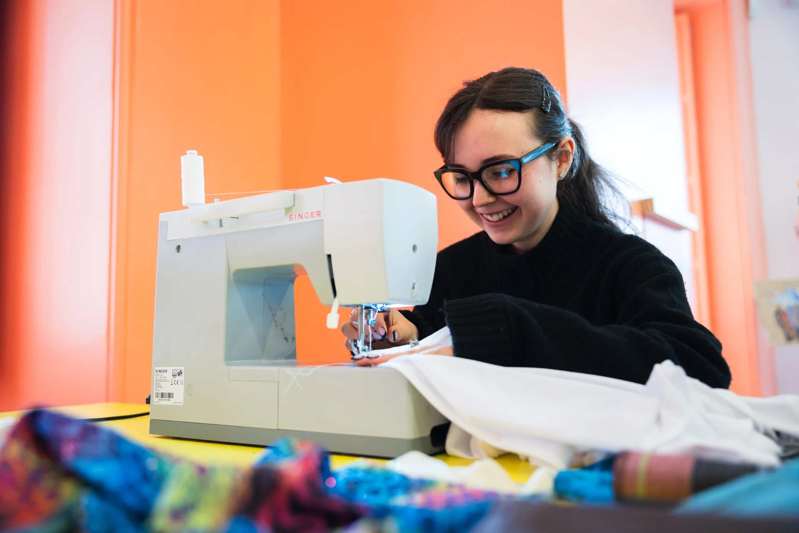 A person sits at a table using a white Singer sewing machine, guiding a piece of white fabric under the needle. The table is covered with colourful fabric pieces in the foreground. The scene is set in a bright indoor workshop space with orange walls and soft natural light coming from windows behind. The sewing machine, fabric, and tools are the main focus of the image.