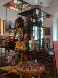 A tall glass-and-metal display structure filled with historic decorative objects arranged across multiple shelves inside a museum gallery. Items include gilded clocks, ceramic vessels, carved wooden furniture panels, sculptural ornaments, and patterned boxes. A richly decorated gold clock and several painted vases are prominent at mid level. Framed paintings hang on the gallery walls behind the case, and daylight enters through a tall arched window on the right, illuminating the objects.