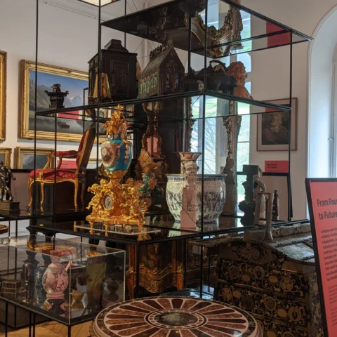 A tall glass-and-metal display structure filled with historic decorative objects arranged across multiple shelves inside a museum gallery. Items include gilded clocks, ceramic vessels, carved wooden furniture panels, sculptural ornaments, and patterned boxes. A richly decorated gold clock and several painted vases are prominent at mid level. Framed paintings hang on the gallery walls behind the case, and daylight enters through a tall arched window on the right, illuminating the objects.