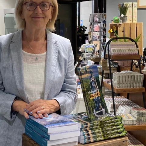 A person stands behind a wooden display table in a museum or gallery shop. Stacks of books are arranged neatly on the table and in wooden crates, with a central display featuring a book titled “Gibside” showing a photograph of a formal landscape on the cover. Behind the table, shelves hold postcards, stationery, and small gift items. The setting includes an arched doorway and soft indoor lighting, suggesting a heritage or museum shop environment.