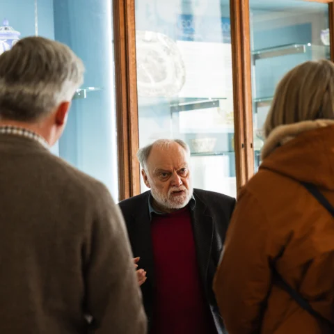A small group of people stand in front of glass display cases in a museum gallery while one person speaks, with ceramics and labelled objects visible behind the glass.