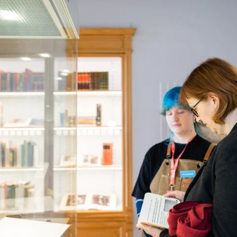 Two people stand beside a glass display case in a museum setting, looking at a book or printed material together, with shelves of books and archival items visible in the background.