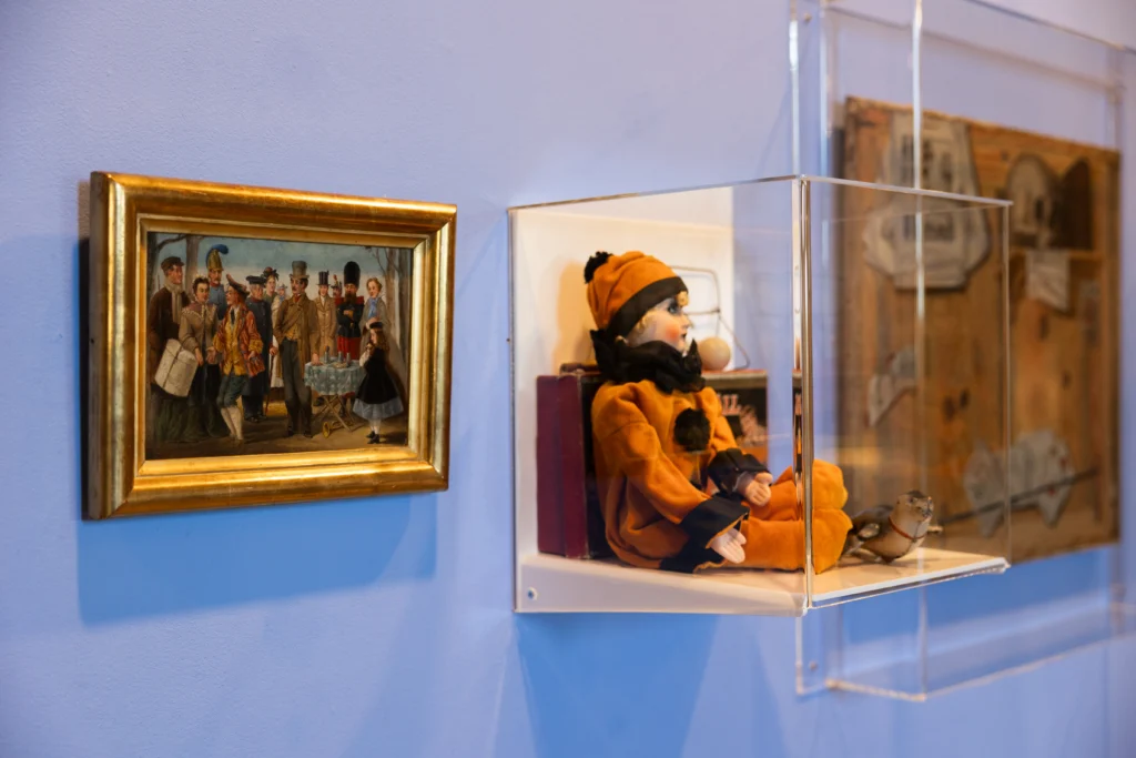 Photograph of a museum display featuring a framed historical painting and a glass case containing a vintage doll dressed in orange with a small bird figurine. The painting depicts a group of people in period clothing, while the display highlights cultural artifacts from different eras with clear lighting and wall-mounted arrangements.