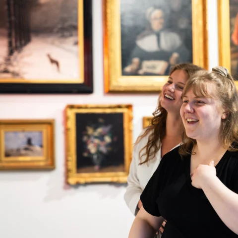 Two visitors stand close together in a gallery, looking at artworks on a wall filled with framed paintings, including a mix of portraits and landscapes.