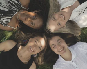 Four people lie on grass with their heads close together in a circular arrangement, photographed from above, with sunlight casting soft shadows across their faces and clothing.