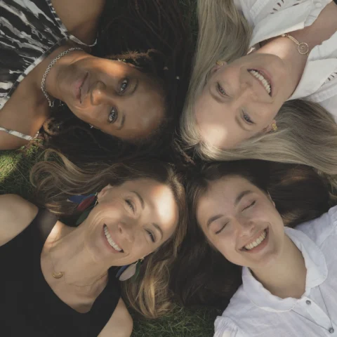 Four people lie on grass with their heads close together in a circular arrangement, photographed from above, with sunlight casting soft shadows across their faces and clothing.