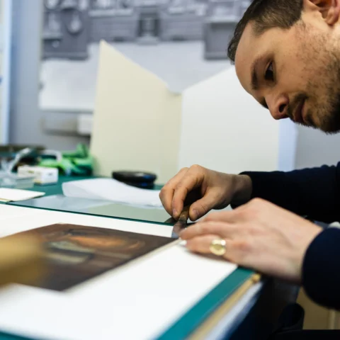 A person leans over a workbench using a small tool to carefully adjust or clean the surface of an artwork. Papers, tools and materials are arranged across the table, with studio equipment and shelving visible in the background.