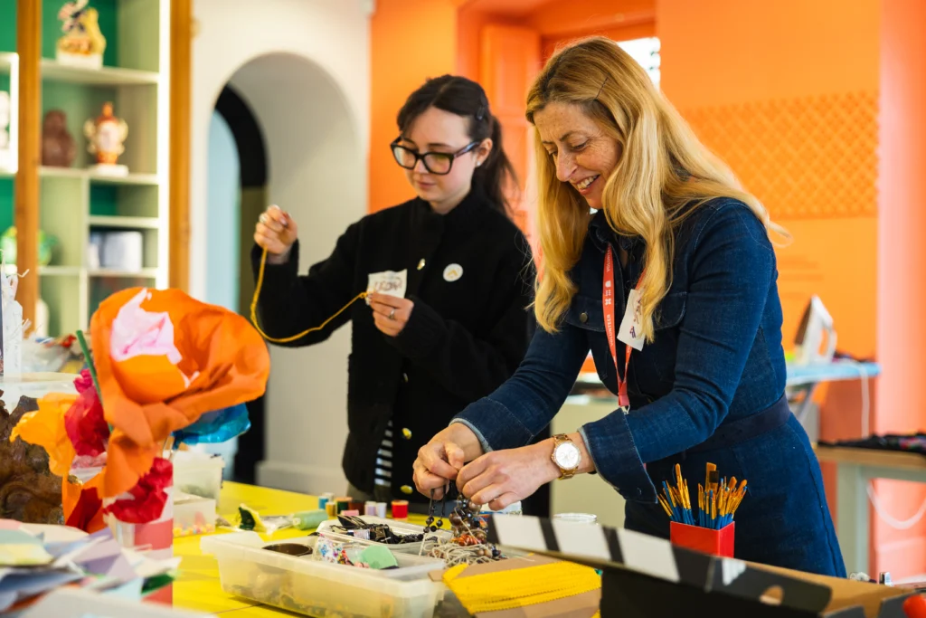 Photograph of two women engaging in a craft activity at a table filled with art supplies, including paper flowers, beads, and paintbrushes. Brightly coloured room with orange walls and shelves in background, highlighting a creative and collaborative environment.