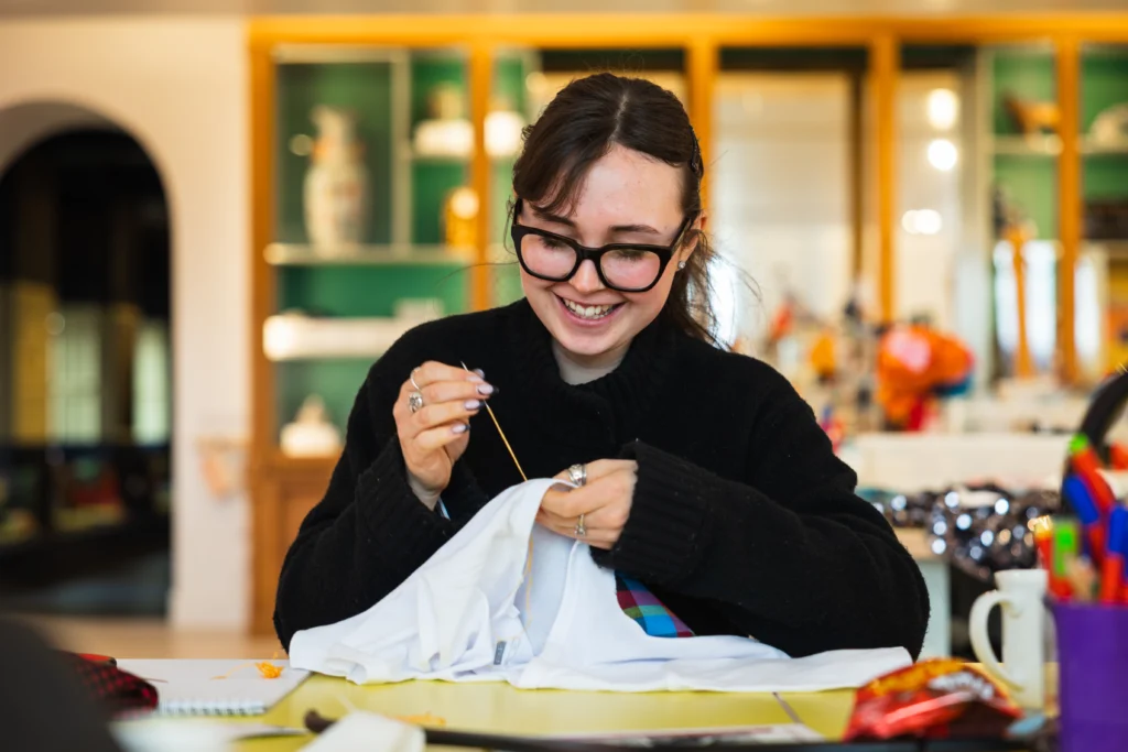 Photograph of a person sewing a white garment using a needle and thread at a table filled with various sewing materials. Background includes shelves with decorative items and a brightly lit room, highlighting a creative workspace environment.