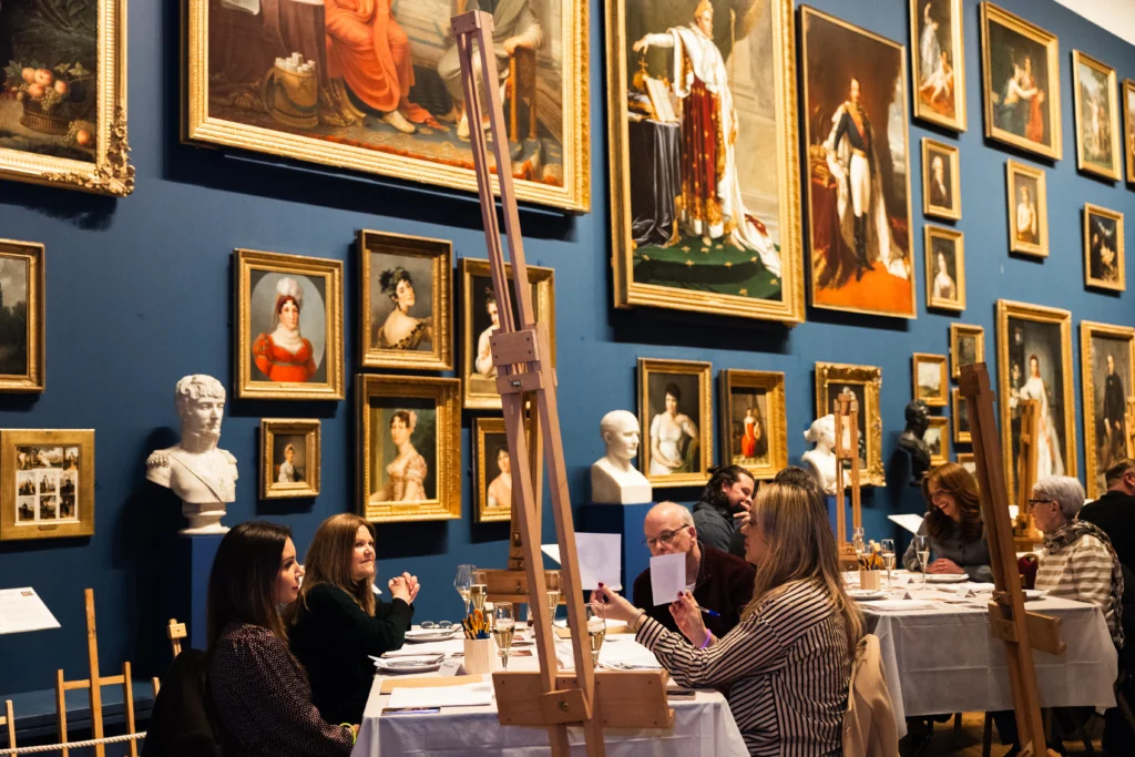 Photograph of an art gallery room with people seated at tables engaged in painting or drawing activities. Walls are adorned with numerous framed classical portraits and bust sculptures, creating an immersive artistic environment.