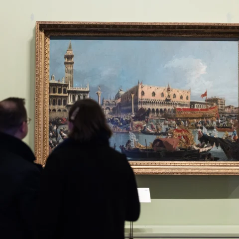 Two visitors stand in a gallery facing a large framed painting of a Venetian waterfront scene with boats and a grand building, surrounded by smaller framed portraits on a pale green wall.