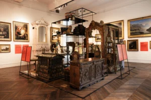 A museum gallery display features ornate antique furniture, ceramics, and decorative objects arranged within a central glass shelving structure, surrounded by framed paintings on white walls and informational panels on stands.