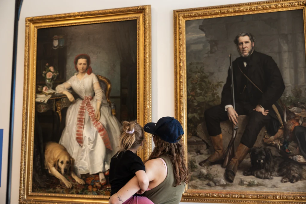 Photograph showing two large framed portraits displayed in a gallery, featuring a woman in a white dress with a dog and a man in dark clothing with hunting dogs. A woman holding a child observes the paintings, highlighting interaction between viewers and classical art pieces with ornate gold frames.