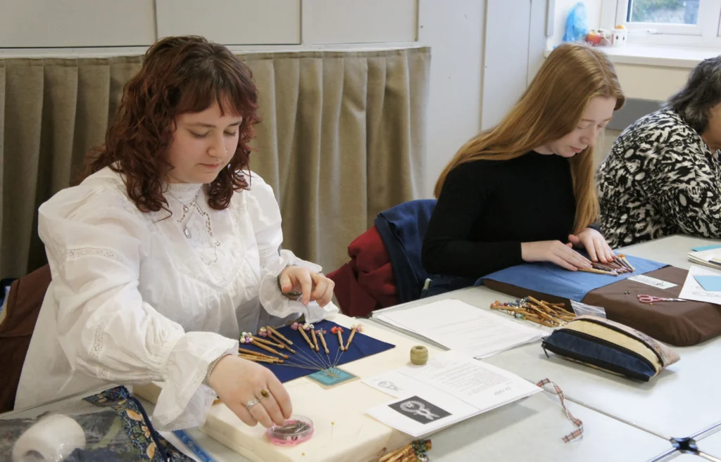 Two people sit at a table working on lace-making, arranging bobbins and thread on blue fabric patterns, with tools, printed instructions, and sewing materials spread across the table in a bright indoor setting.
