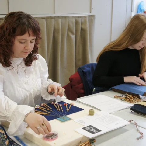 Two people sit at a table working on lace-making, arranging bobbins and thread on blue fabric patterns, with tools, printed instructions, and sewing materials spread across the table in a bright indoor setting.
